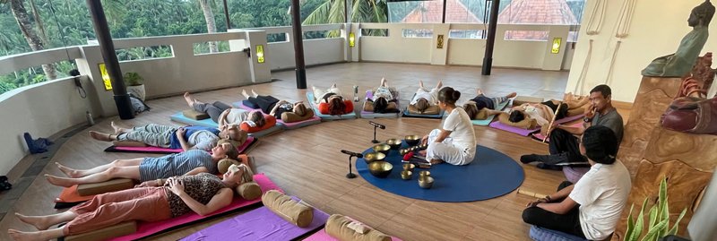 a group of 11 people lay in a semi circle with their heads facing in towards the center. A Balinese woman dressed in white sits at the center surrounded by brass Singing Bowls. Two flute musicians sit behind her.